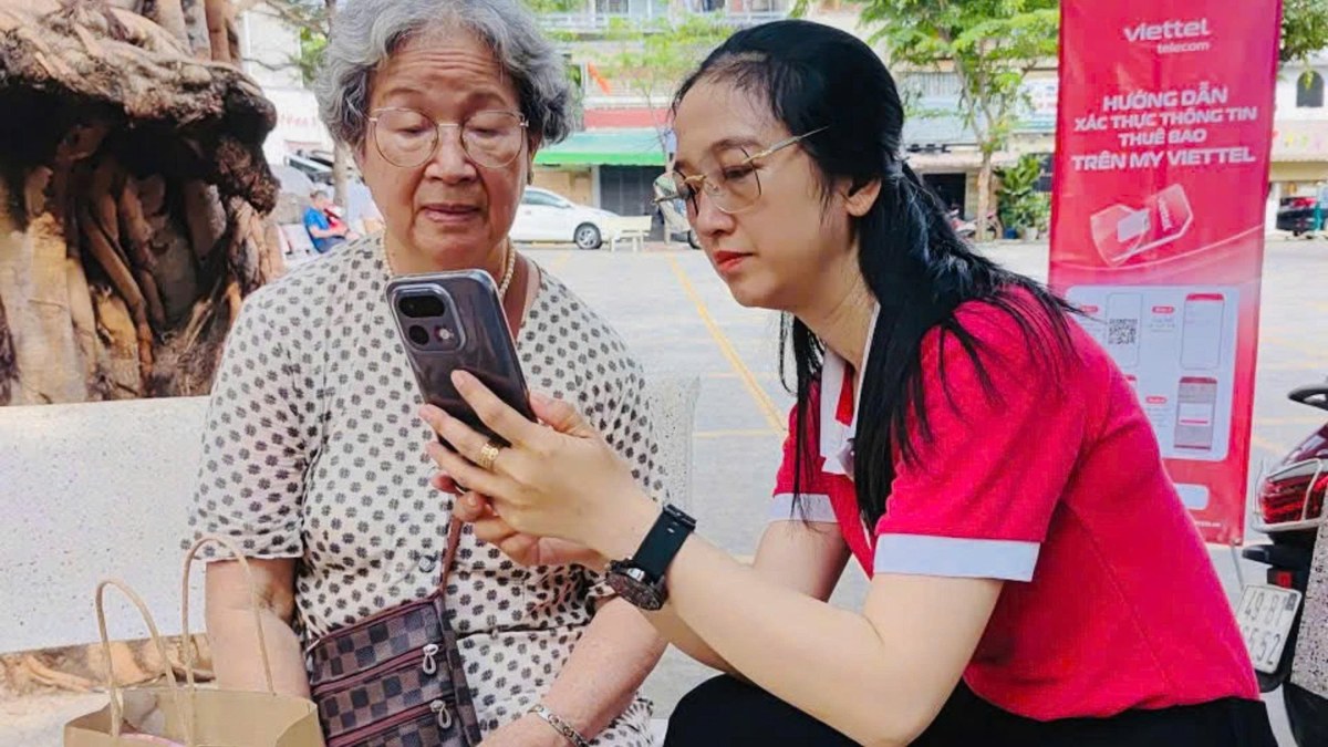 Cô gái áo đỏ hướng dẫn cụ bà tóc bạc dùng điện thoại cạnh banner Viettel ngoài trời.